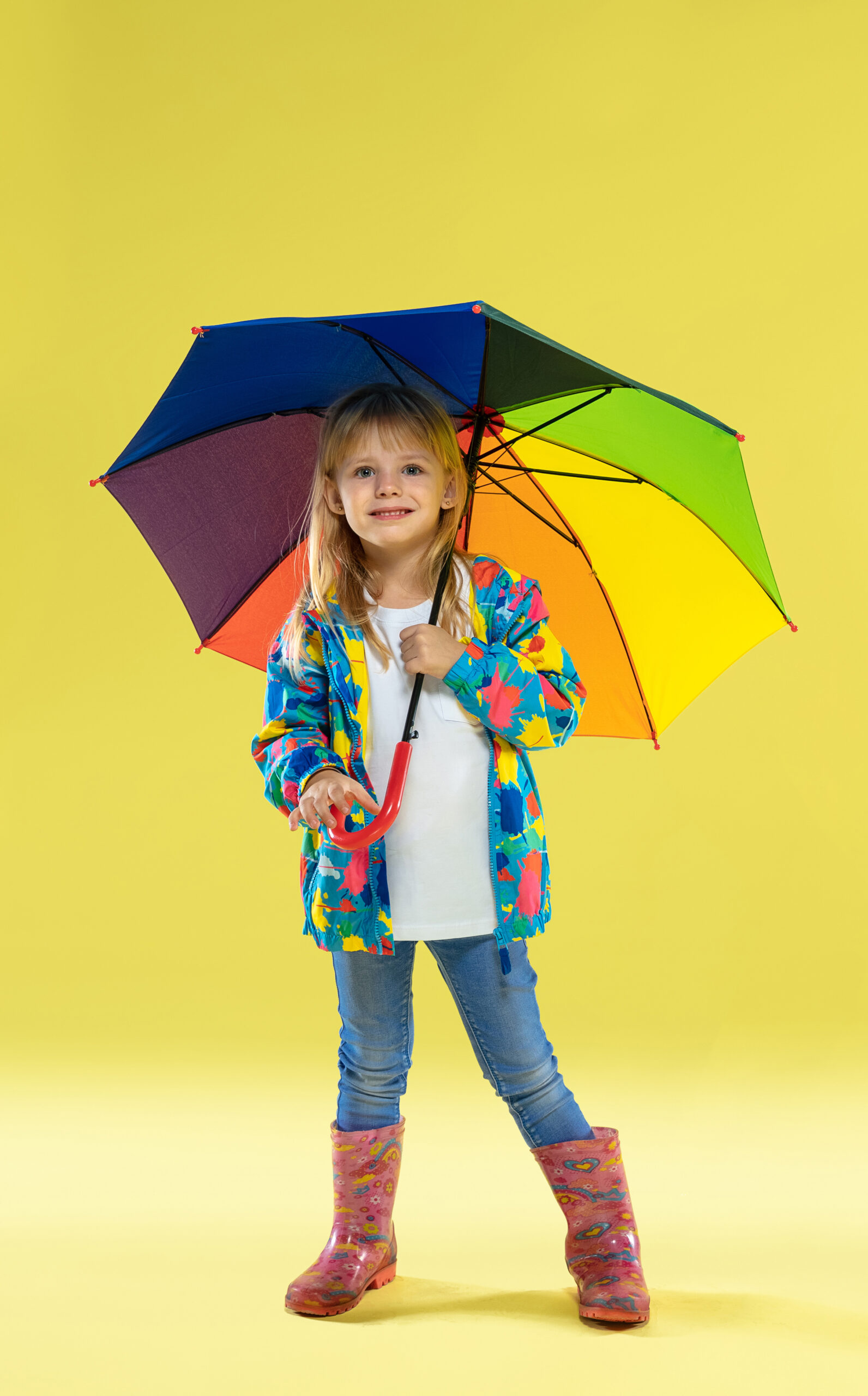 A full length portrait of a bright fashionable girl in a raincoat a full length portrait of a bright fashionable girl in a raincoat holding an umbrella of rainbow colors on yellow studio background scaled 1
